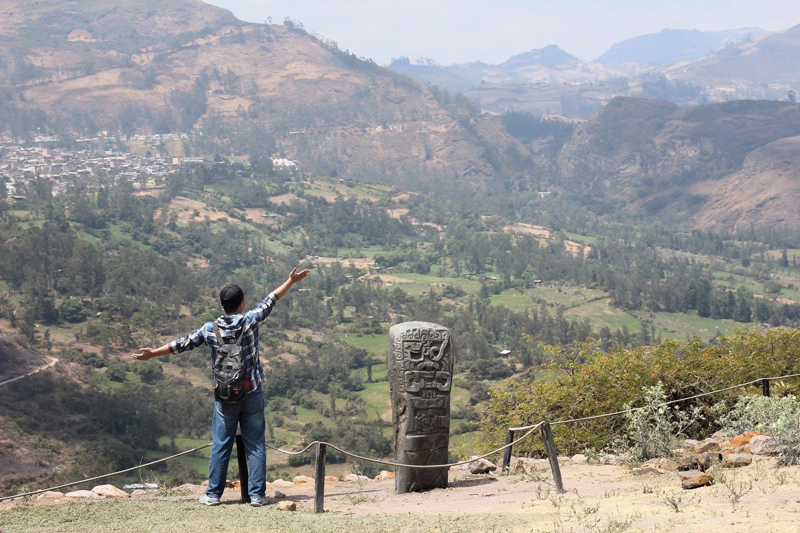 A visitor with arms outstretched beside a carved monolith at Cuntur Wasi, the valley falling away behind
