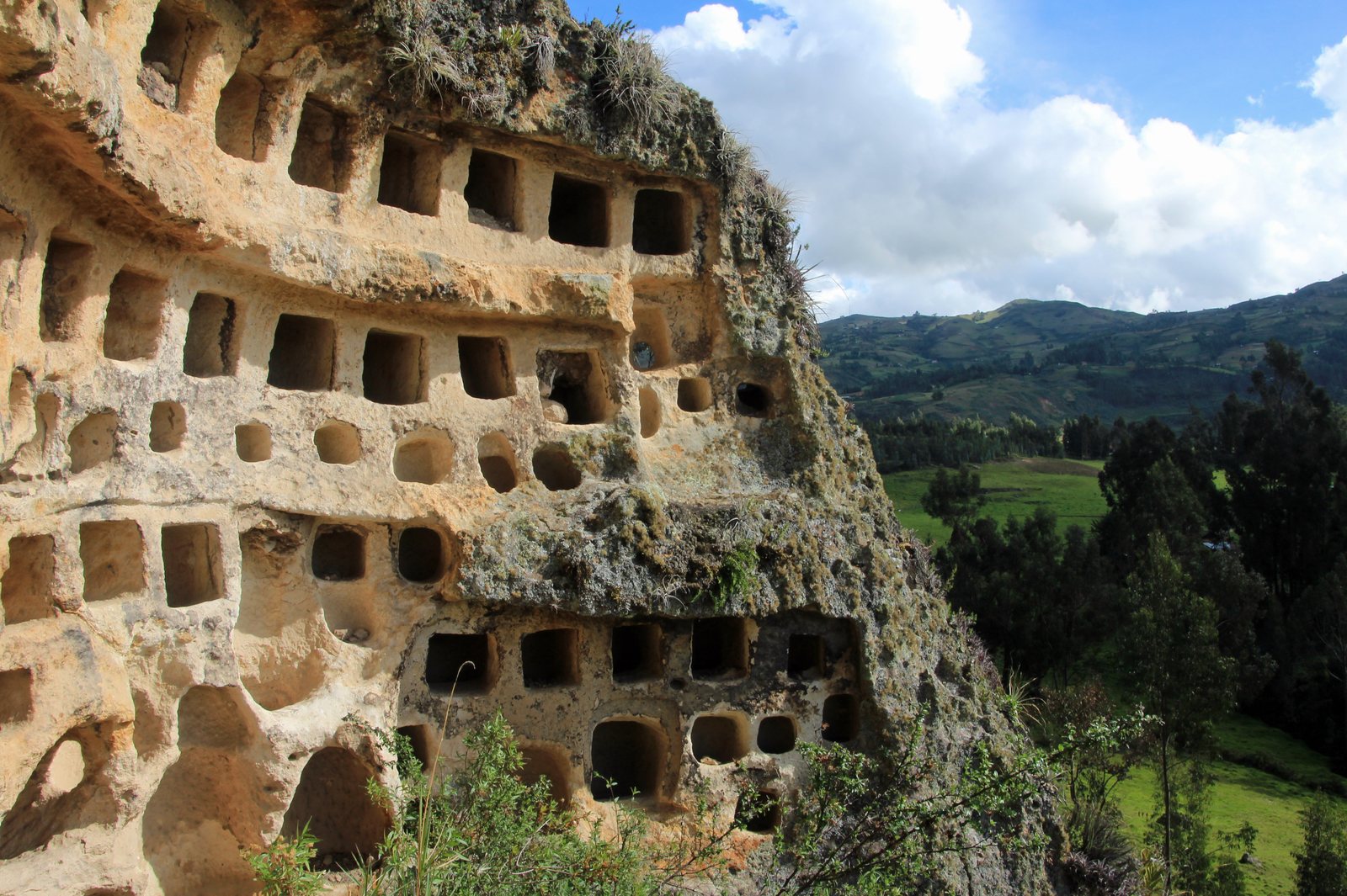 Rows of square burial niches cut into the volcanic cliff face at Otuzco, with farmland in the valley below